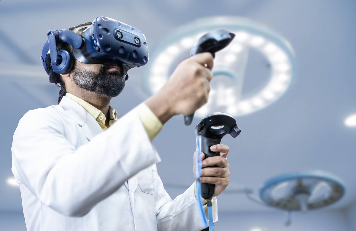A clinician in a white coat wearing a VR headset and holding two motion controllers under operating theatre lights.